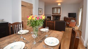 A dining room with a wooden table set for four, chairs, a vase with flowers, a fireplace, and a television in the background. - 2 Bed in Penmaenmawr (oc-56081) (Penmaenmawr)