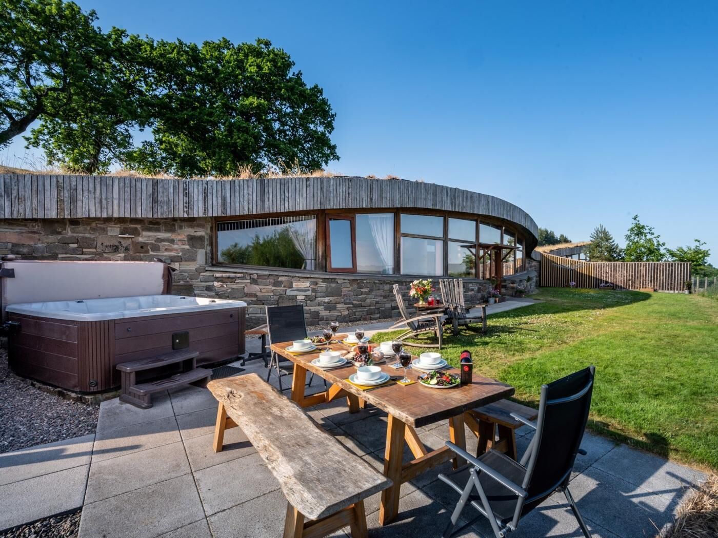 A curved stone building with a hot tub, a wooden table set for dining, and a grassy yard.