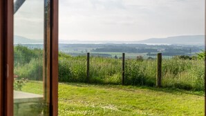 Une vue à travers une fenêtre à cadre en bois, sur un champ verdoyant, une clôture et des collines lointaines.