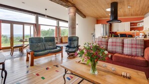 Un vaste espace de vie doté d’un plancher en bois, de deux canapés en cuir, d’une table basse ornée d’un vase de fleurs, et d’une cuisine en arrière-plan.