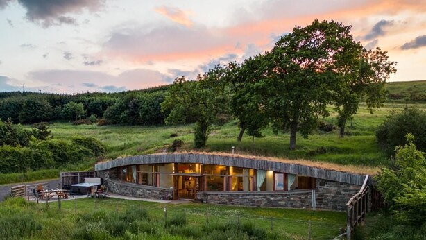 Un bâtiment moderne en pierre, de forme circulaire et surmonté d’un toit de chaume, entouré d’une végétation luxuriante et d’arbres.