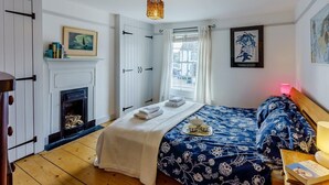 A bedroom with a bed, a fireplace, a window with curtains, and a painting on the wall. - 3 Bed in Whitstable (oc-wcc12) (Whitstable)