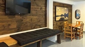 Una sala de estar moderna con televisor de pantalla plana, paneles de madera, un banco, una mesa de comedor con sillas y un cuadro enmarcado en la pared.