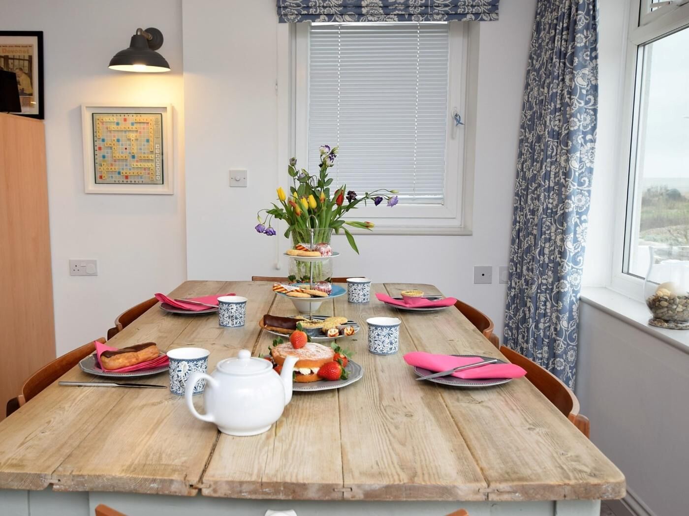 A wooden dining table with a white teapot, pink napkins, and a floral centerpiece.