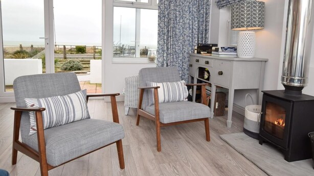 Una sala de estar acogedora con dos sillones grises, un aparador blanco con adornos, una chimenea y vistas al exterior.
