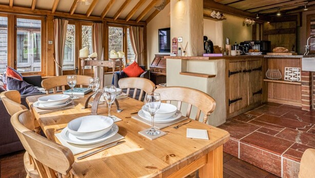 Una mesa de comedor de madera con platos, vasos y cubiertos, una zona de cocina con barra, y un rincón de estar acogedor con sofá y sillones.