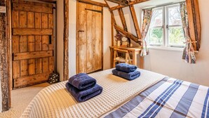 Un dormitorio con vigas de madera a la vista, un ventanal con cortinas florales y una cama con colcha a rayas y toallas dobladas.