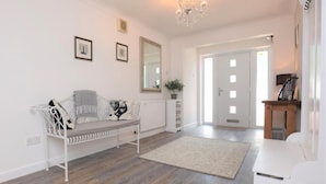A modern living room with a white sofa, a white bench, a chandelier, and a door leading to another room. - 3 Bed in Dereham (oc-56661) (Horningtoft)