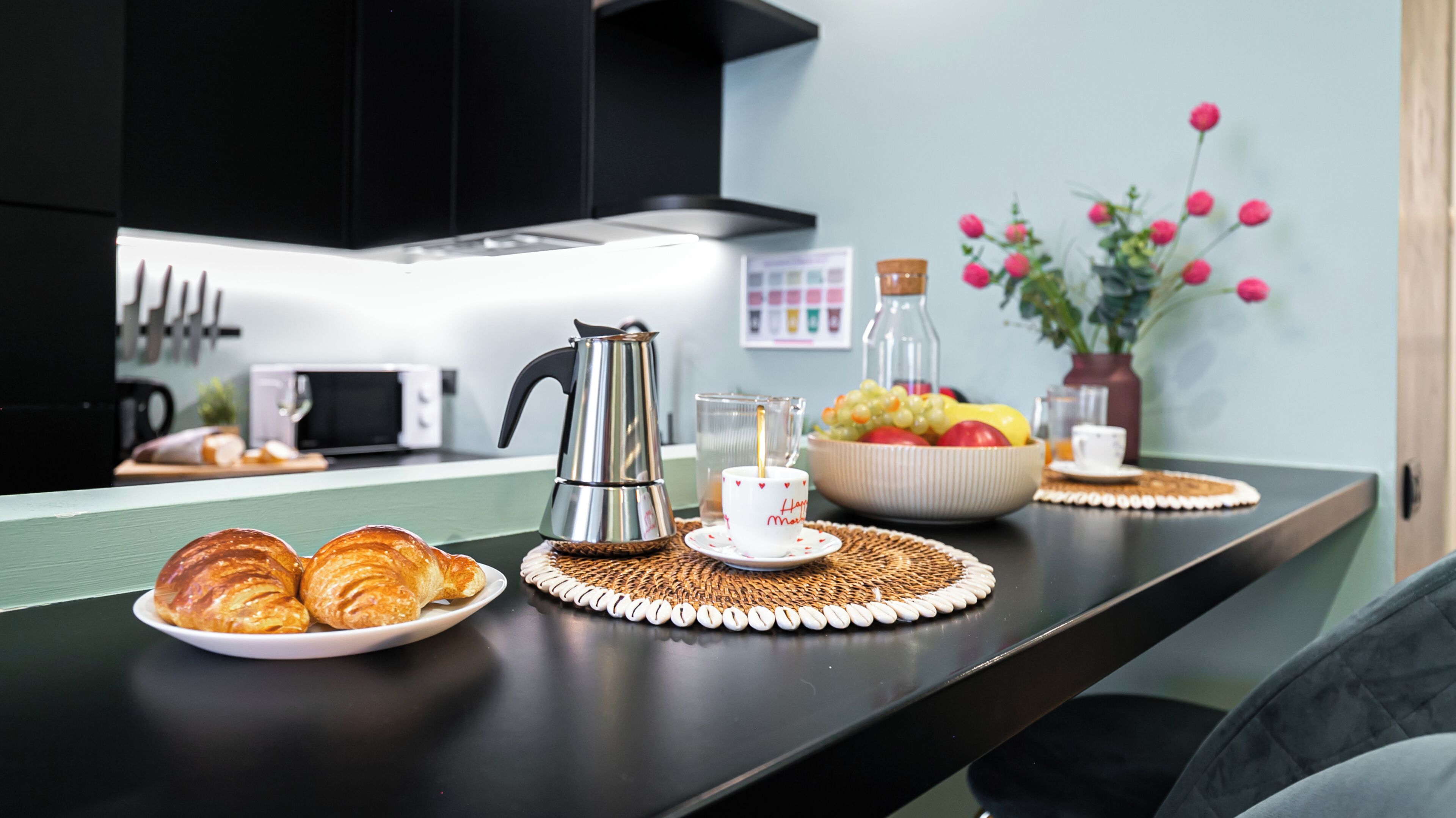 A kitchen counter with croissants, a coffee pot, a fruit bowl, and a vase with flowers.