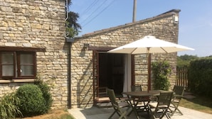 Ein Steinhaus mit einer Terrasse, auf der sich ein Tisch und Stühle befinden, eine offene Tür und ein weißer Sonnenschirm.