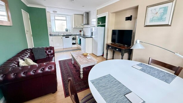 A compact living space with a dining area, a sofa, a television, and a kitchen in the background. - Alway at Lifton Hall (Lifton)