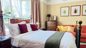 A bedroom with a bed, a nightstand, a dresser, and a red sofa. There are curtains, a lamp, and framed botanical prints on the wall. - Alway at Lifton Hall (Lifton)