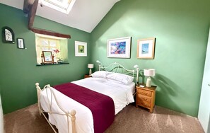 A bedroom with a bed, nightstands, a window with blinds, and wall-mounted pictures. - The Barn at Lifton Hall (Lifton)
