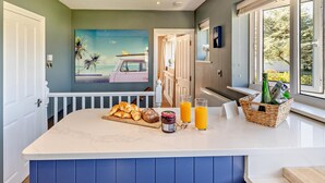 A kitchen with a marble countertop, a painting of a van and palm trees, and a window with a view of trees. - 4 Bed in Braunton (oc-56111) (Croyde)