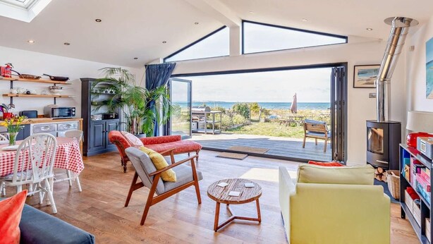 Uma sala de estar espaçosa com cozinha, área de jantar e uma grande porta de vidro deslizante que dá para uma varanda com vista para a praia.