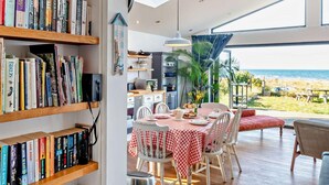 Cozinha moderna com vista para a praia, um espaço de refeições com toalha quadriculada e uma estante cheia de livros.