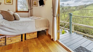 Een slaapkamer met een bed, houten vloer, gordijnen, een televisie en uitzicht op een groen landschap.