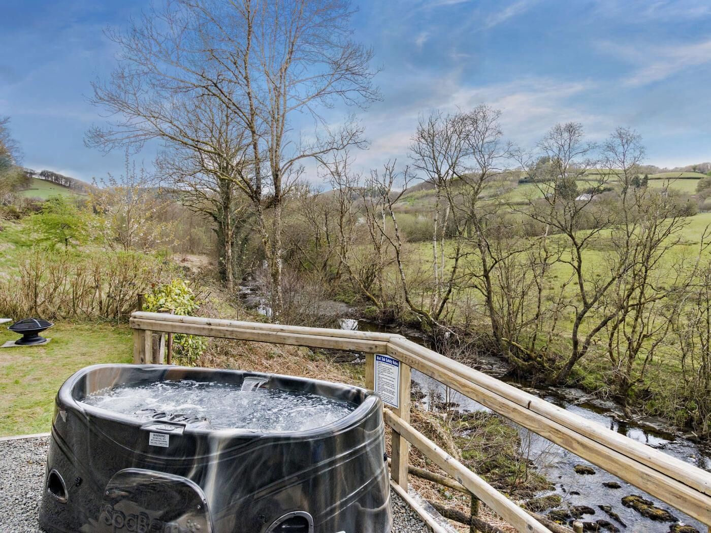Un bain à remous doté d’une balustrade en bois, entouré d’un paysage naturel avec des arbres et un ruisseau.