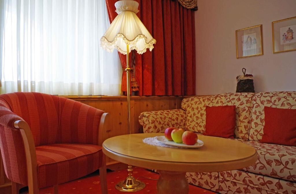 A room with a striped armchair, a sofa with a floral pattern, a round table with a fruit plate, a floor lamp, and a window with curtains.