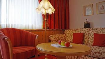 A room with a striped armchair, a sofa with a floral pattern, a round table with a fruit plate, a floor lamp, and a window with curtains.