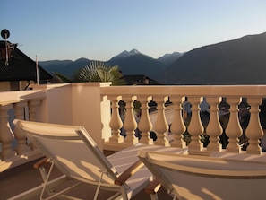 A balcony with two white lounge chairs, a potted plant, and a view of distant mountains.