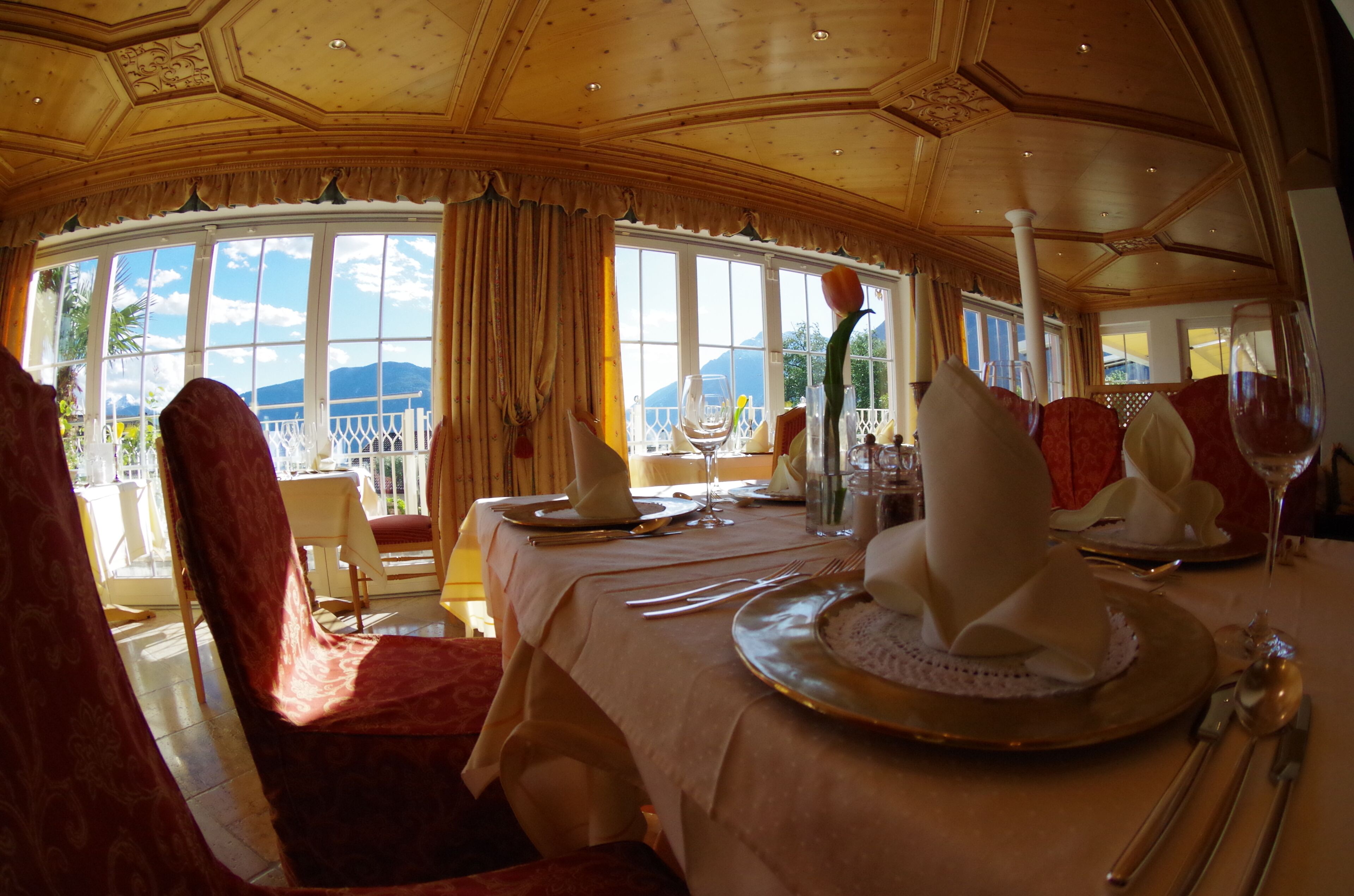 A dining area with a table set for a meal, large windows with a view of mountains, and a high ceiling with ornate woodwork.