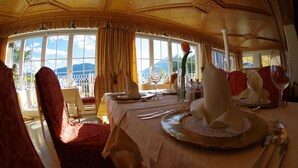 A dining area with a table set for a meal, large windows with a view of mountains, and a high ceiling with ornate woodwork. - Hotel Bergland (Schenna)