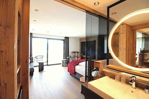 A modern hotel room with a large bed, a round mirror, a bathroom with a sink, and a view of the outside.