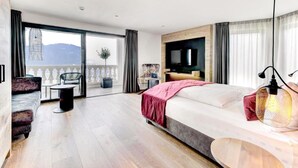 A modern hotel room with a large bed, a TV, a balcony with a view, and a seating area with a chair and table. - Hotel Bergland (Schenna)
