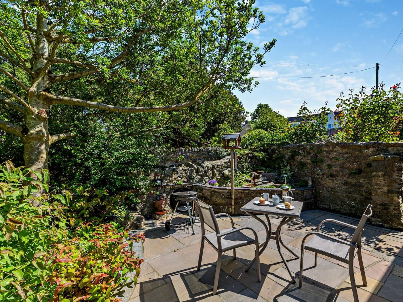 An outdoor patio with a table and chairs, surrounded by greenery and a stone wall.