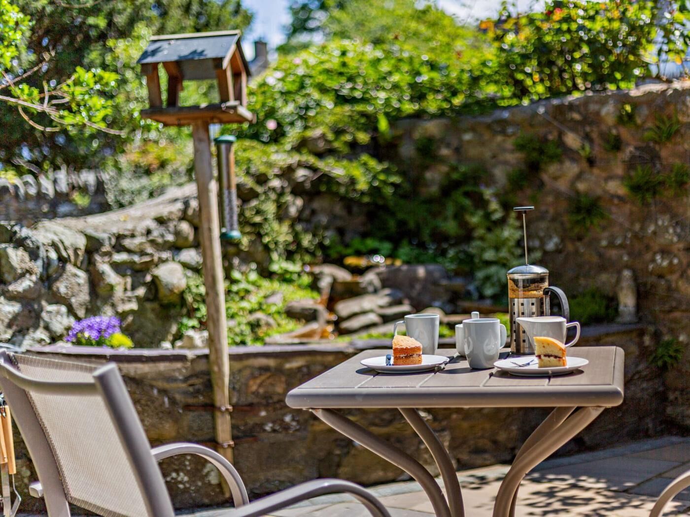 Un tavolo da esterno con una caffettiera, tazze e una fetta di torta, sullo sfondo del verde e un muro in pietra.