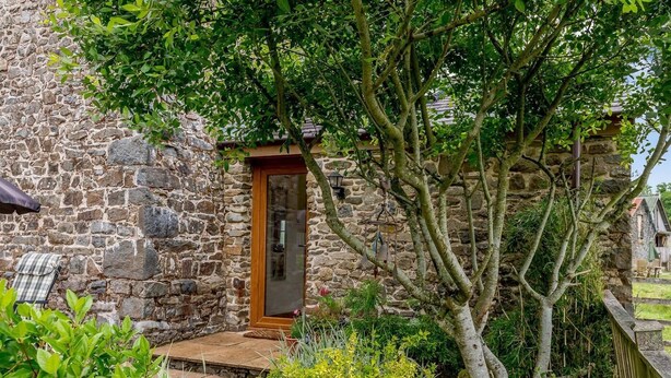 Un edificio de piedra con puerta de madera, un pequeño porche y abundante vegetación alrededor.
