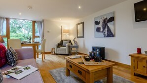Una sala de estar acogedora con un sofá, una mesa de centro y un televisor. Se ve una ventana con cortinas, una lámpara y un cuadro de una vaca.