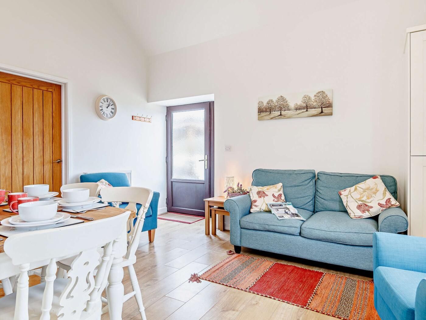 Una sala de estar con un sofá azul, una mesa de comedor con sillas, un reloj en la pared y una puerta que da al exterior.