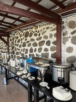 A buffet setup with various food containers and utensils on a table, set against a stone wall.