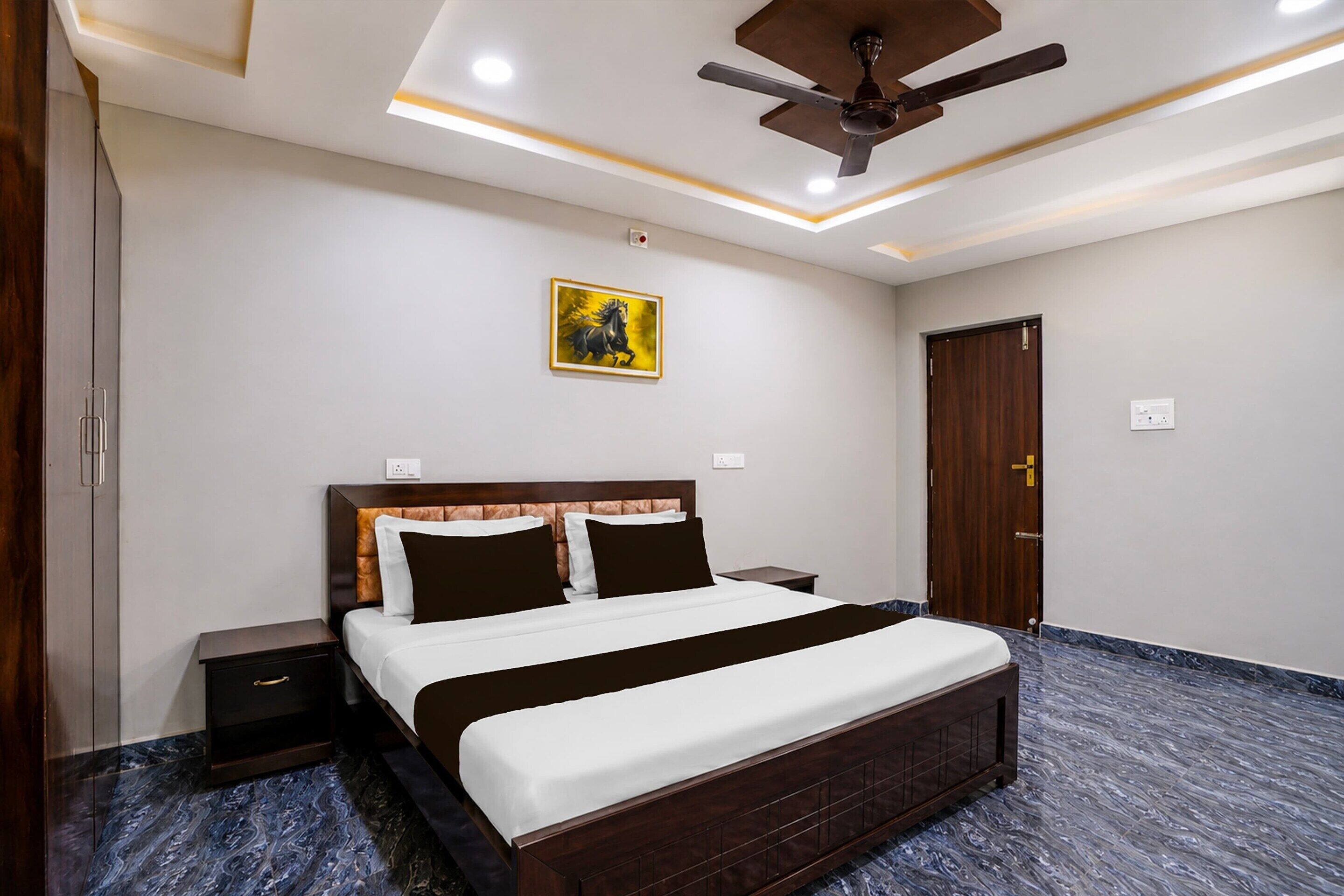 Una habitación de hotel con cama, mesitas de noche, ventilador de techo, un cuadro y una puerta.