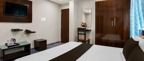 A hotel room with a bed, bedside table, a mirror, a wardrobe, a television mounted on the wall, and a small desk with a telephone and a tray with cups and a kettle.