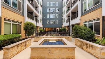 A multi-story residential building with balconies, a central courtyard with a water feature, and a stone pathway.