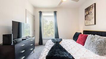 Un dormitorio con cama, cómoda, televisión, ventilador de techo, ventana con cortinas y un cuadro en la pared.