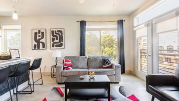 A modern living room with a sofa, coffee table, and chairs, a dining area with a bar, and large windows with curtains.