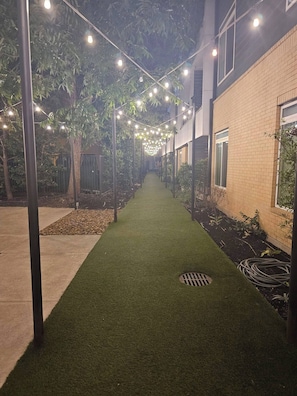 Camí amb gespa artificial, envoltat d'arbres i edificis, sota llums de garlandes.