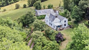 A white house with a large porch and a covered patio, surrounded by lush greenery and hills. - 3 Bed in Crickhowell (oc-56811) (Llangynidr)