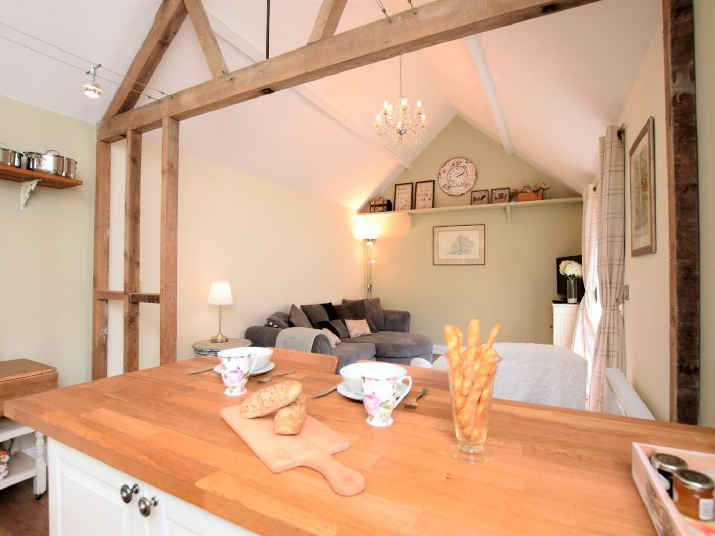 Eine gemütliche Küche mit Holzbalken, einem Esstisch mit Brot und Tassen sowie einer Wohnbereiches mit Sofa und Wanddekoration.