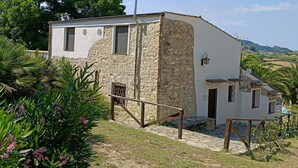 Un edificio rústico de piedra con fachada blanca, una escalera de madera que lleva a una puerta y un pequeño jardín con plantas tropicales.