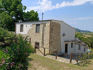 Ein rustikales Steinhaus mit weißer Fassade, einer Holztreppe, die zu einer Tür führt, und einem kleinen Gartenbereich mit tropischen Pflanzen.