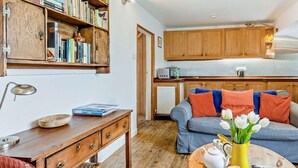 Ein gemütliches Wohnzimmer mit einem Holztisch, einem Sofa mit orangefarbenen Kissen, einem Couchtisch mit Teekanne und Blumen sowie einem Bücherregal voller Bücher.