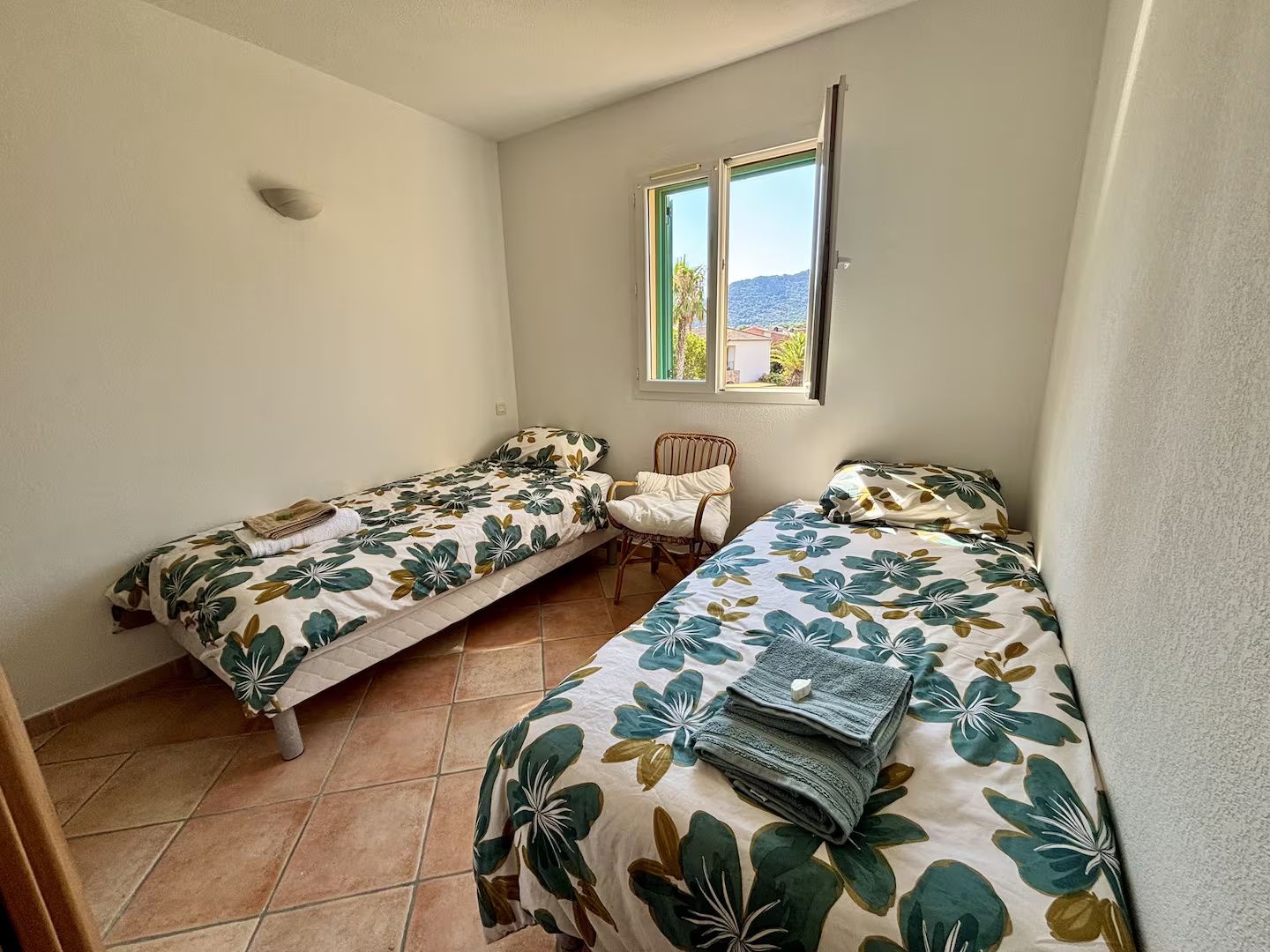 A small, single-room accommodation with two single beds, a chair, and a window providing a view of the outdoors.