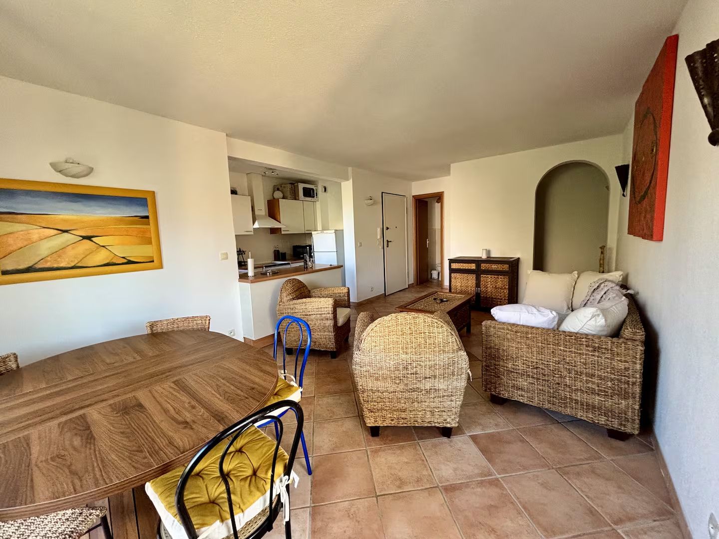 A living room with a dining table, wicker furniture, and a kitchen in the background.
