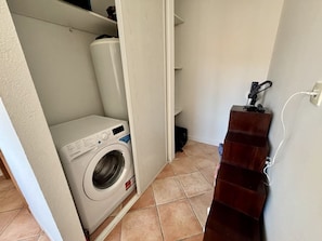 Une petite buanderie équipée d’un lave-linge à chargement frontal, d’une étagère en bois et d’un sol carrelé.
