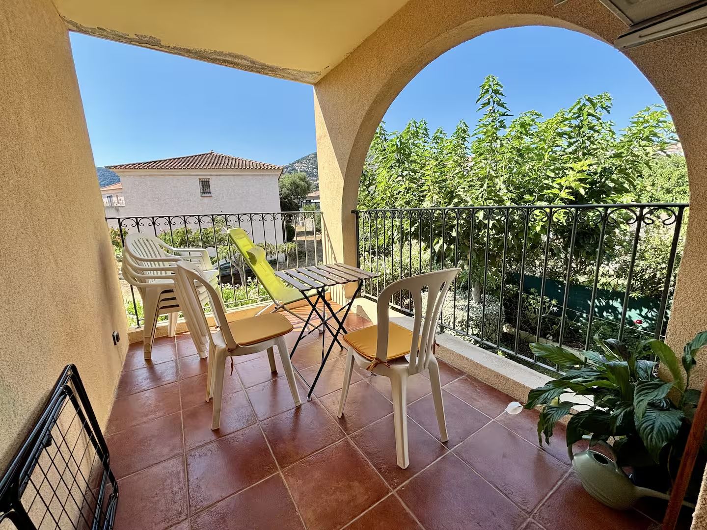 Un balcon avec une table et des chaises, un pot de fleurs et une vue sur un immeuble et des arbres.